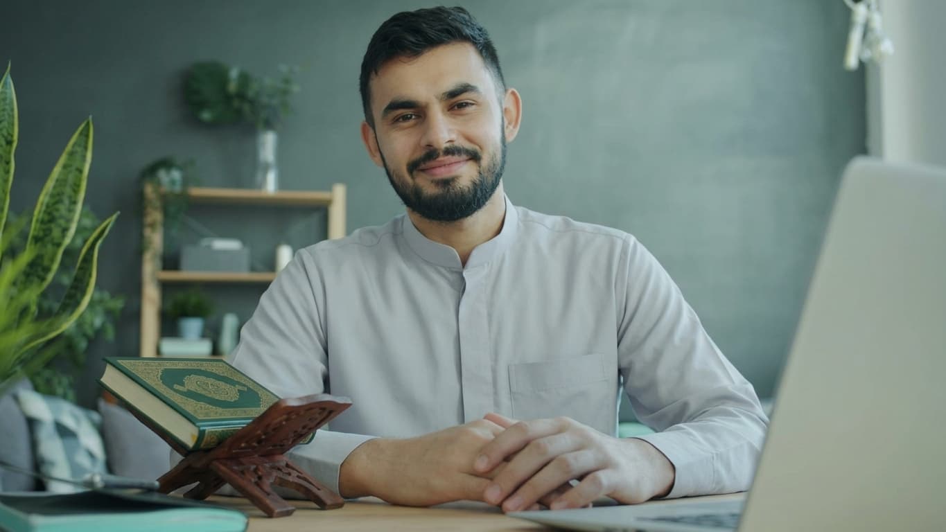 Smiling male Quran teacher at his desk with a Quran on a stand, ready for an online lesson